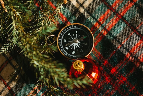 A compass is placed on a plaid fabric with shades of green, red, and brown. To the bottom right of the compass is a shiny red Christmas ornament with a golden top. Pine needles are visible around the compass, adding a festive touch.