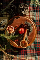 A cake decorated with dried orange slices, cinnamon sticks, rosemary sprigs, and star anise is placed on a plaid cloth. The scene includes pinecones and festive decorations, giving a rustic and cozy vibe.