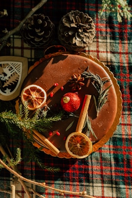 A cake decorated with dried orange slices, cinnamon sticks, rosemary sprigs, and star anise is placed on a plaid cloth. The scene includes pinecones and festive decorations, giving a rustic and cozy vibe.