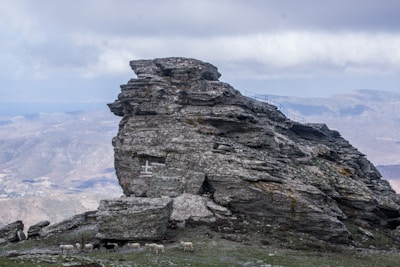 A large rocky formation occupies the center with rugged textures and a cross symbol painted on it. Below the rock, a small group of sheep graze on sparse grass. The background reveals a mountainous landscape under a cloudy sky, providing a dramatic backdrop.