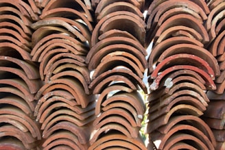 A stack of old, curved terracotta roof tiles with a rustic appearance, showing signs of weathering and slight damage. The tiles are uniformly arranged in rows, creating a repetitive pattern and texture.