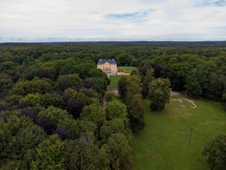 Aerial view of a large yellow building with a blue roof nestled among dense green forests and expansive lawns. A long, tree-lined path leads to the front entrance of the building. The landscape stretches out into the horizon under a partly cloudy sky.
