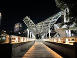 A modern bridge with a geometric steel framework is illuminated by artificial lights at night. The pathway leading to the bridge is wooden, flanked by white railings. Tall buildings with lit windows form the background, and trees are visible to the right.