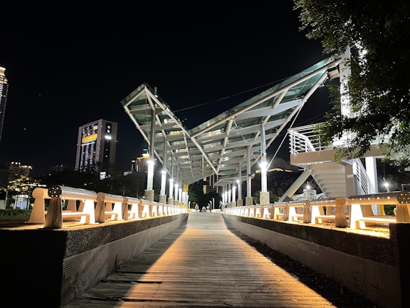 A modern bridge with a geometric steel framework is illuminated by artificial lights at night. The pathway leading to the bridge is wooden, flanked by white railings. Tall buildings with lit windows form the background, and trees are visible to the right.