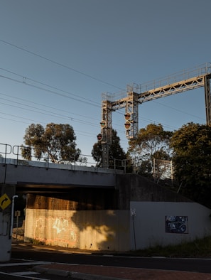 A railway bridge structure with multiple signal lights is set against a clear sky. In the foreground, there are trees and shadowed concrete walls, one of which is adorned with graffiti. Metal railings line the bridge, and power lines stretch across the scene.