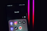 A smartphone screen displays a folder labeled 'Social,' containing icons for various social media apps, including Facebook, Instagram, Threads, X, LinkedIn, TikTok, and YouTube. The background is dark with pink and red neon lights.