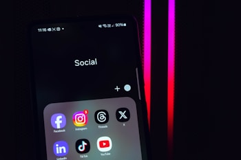 A smartphone screen displays a folder labeled 'Social,' containing icons for various social media apps, including Facebook, Instagram, Threads, X, LinkedIn, TikTok, and YouTube. The background is dark with pink and red neon lights.