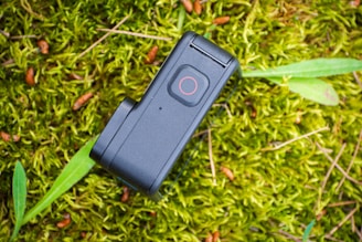 A small, rectangular black action camera with a visible power button is placed on a vibrant green mossy surface. Some small brown seed cones and green leaves are scattered around the camera, adding texture to the natural setting.