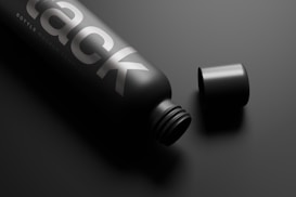 A sleek, black matte bottle lies on a dark surface with the cap placed next to it. The bottle features bold silver text with a minimalist design, creating a sophisticated and modern look.