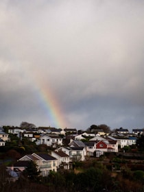 A residential area with multiple houses and lush greenery, featuring a vivid rainbow emerging from a cloudy sky in the background.