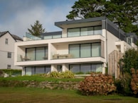 A modern three-story house featuring large glass windows and flat roofs, surrounded by lush greenery and a neatly manicured lawn. The structure includes outdoor seating areas on the terraces. The sky above appears partly cloudy, and a neighboring house is visible on the left side.