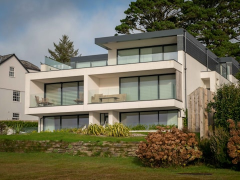 A modern three-story house featuring large glass windows and flat roofs, surrounded by lush greenery and a neatly manicured lawn. The structure includes outdoor seating areas on the terraces. The sky above appears partly cloudy, and a neighboring house is visible on the left side.