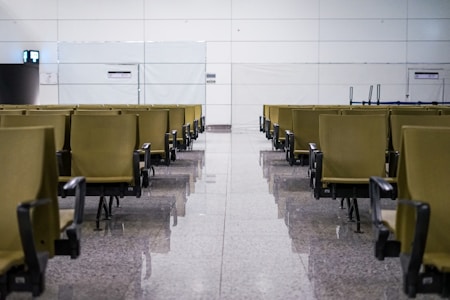 A row of empty waiting chairs is arranged in a symmetrical pattern in a brightly lit, modern indoor space with a reflective floor. The wall in the background is paneled and minimalistic, with two small signs or labels visible. The lighting is even, creating a clean and orderly atmosphere.