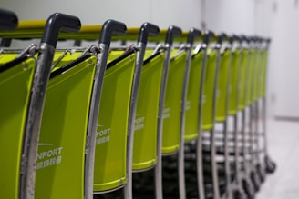 A line of green luggage carts with black handles arranged neatly in a row, positioned in an indoor setting. The background appears to be a plain wall and floor, suggesting an airport or transportation facility.