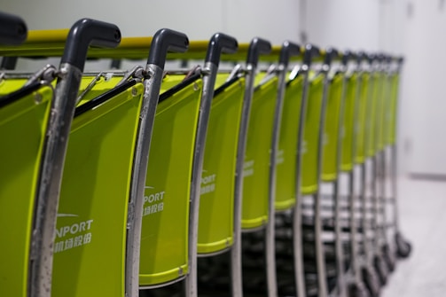 A line of green luggage carts with black handles arranged neatly in a row, positioned in an indoor setting. The background appears to be a plain wall and floor, suggesting an airport or transportation facility.