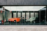 Two vintage vehicles, an orange convertible car and a green classic sedan, are displayed behind a large glass window. A motorcycle is positioned between them. The interior is spacious and modern, with a polished floor reflecting the vehicles, suggesting a showroom setting.