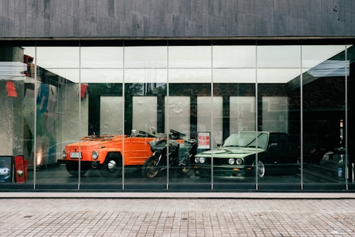 Two vintage vehicles, an orange convertible car and a green classic sedan, are displayed behind a large glass window. A motorcycle is positioned between them. The interior is spacious and modern, with a polished floor reflecting the vehicles, suggesting a showroom setting.