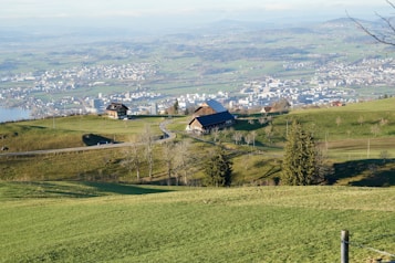 A serene landscape featuring rolling green hills with a rural farmhouse and a barn nestled among them. The scene overlooks a sprawling cityscape in the distance with clear skies and a body of water visible. Trees and winding roads are scattered throughout the countryside, adding to the tranquil nature of the setting.