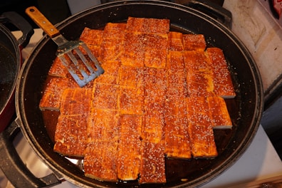 A large frying pan filled with evenly arranged rectangular tofu pieces, coated with a reddish sauce and sprinkled with sesame seeds. A metal spatula with a wooden handle is resting against a few pieces of tofu on the left side of the pan.