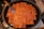 A large frying pan filled with evenly arranged rectangular tofu pieces, coated with a reddish sauce and sprinkled with sesame seeds. A metal spatula with a wooden handle is resting against a few pieces of tofu on the left side of the pan.
