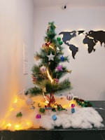 A decorated artificial Christmas tree is adorned with colorful ornaments, including stars, bells, and gift boxes, placed on a bed of artificial snow. The background features a wall-mounted world map silhouette, adding a modern touch to the festive scene.