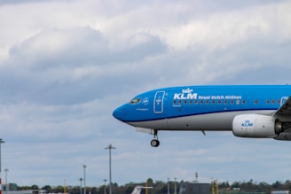 A KLM Royal Dutch Airlines aircraft is in the lower right of the frame, captured in mid-flight against a backdrop of a cloudy sky. The horizon and some airport structures can be seen in the distance, beneath the aircraft.