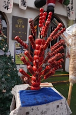 A display of traditional Chinese candied fruit skewers arranged in a decorative manner on what appears to be a market stall. The skewers are covered with shiny, red candy glaze and are displayed in a fan-like arrangement. They are set on a table covered with a white cloth featuring Chinese calligraphy. Behind the display, there are signs with Chinese characters, as well as holiday decorations including a Christmas tree.