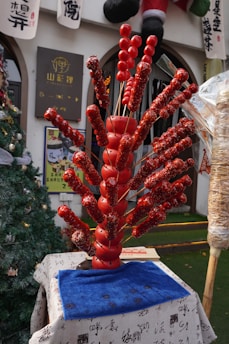 A display of traditional Chinese candied fruit skewers arranged in a decorative manner on what appears to be a market stall. The skewers are covered with shiny, red candy glaze and are displayed in a fan-like arrangement. They are set on a table covered with a white cloth featuring Chinese calligraphy. Behind the display, there are signs with Chinese characters, as well as holiday decorations including a Christmas tree.