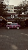 A vintage purple car is parked in front of a restaurant with a rustic exterior, featuring a thatched roof and surrounded by lush greenery. The restaurant has round signage with both English and Hindi fonts above the entrance.