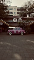 A vintage purple car is parked in front of a restaurant with a rustic exterior, featuring a thatched roof and surrounded by lush greenery. The restaurant has round signage with both English and Hindi fonts above the entrance.