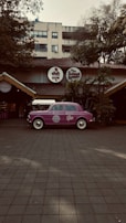 A vintage purple car is parked in front of a restaurant with a rustic exterior, featuring a thatched roof and surrounded by lush greenery. The restaurant has round signage with both English and Hindi fonts above the entrance.
