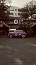 A vintage purple car is parked in front of a restaurant with a rustic exterior, featuring a thatched roof and surrounded by lush greenery. The restaurant has round signage with both English and Hindi fonts above the entrance.
