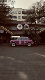 A vintage purple car is parked in front of a restaurant with a rustic exterior, featuring a thatched roof and surrounded by lush greenery. The restaurant has round signage with both English and Hindi fonts above the entrance.
