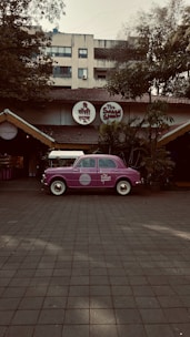 A vintage purple car is parked in front of a restaurant with a rustic exterior, featuring a thatched roof and surrounded by lush greenery. The restaurant has round signage with both English and Hindi fonts above the entrance.