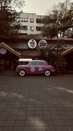 A vintage purple car is parked in front of a restaurant with a rustic exterior, featuring a thatched roof and surrounded by lush greenery. The restaurant has round signage with both English and Hindi fonts above the entrance.
