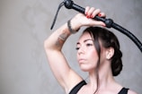 An individual with braided hair holds a black whip above their head. They have visible tattoos on their right arm and red nail polish. The background is a muted gray, adding contrast to the subject's skin and hair.