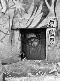 A black and white image featuring graffiti on a wall with a closed, aged metal door. The door has a street art face painted on it, surrounded by abstract mural designs. Several 'love' themed posters are pasted on the wall to the right. The ground is rough with scattered leaves and debris.
