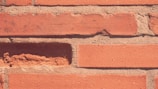 A close-up view of a brick wall with reddish-brown bricks and textured mortar joints. One of the bricks appears partially missing, revealing some rough surfaces underneath.