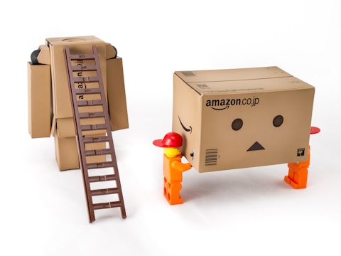 Two toy figures are interacting with a large cardboard box that has the appearance of a robot with a simple face drawn on it. The box stands on four small legs, resembling a character with arms. A brown ladder is placed on another cardboard box figure, leaning against it. The figures appear to be in a playful setting, using everyday objects creatively.