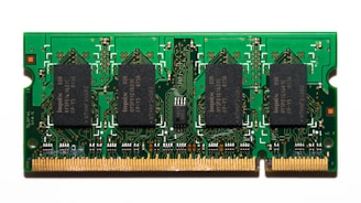 A compact green circuit board features multiple black rectangular chips surrounded by intricate gold pathways and connectors. The detailed design is typical of a RAM module used in computers.