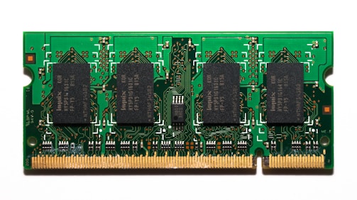 A compact green circuit board features multiple black rectangular chips surrounded by intricate gold pathways and connectors. The detailed design is typical of a RAM module used in computers.