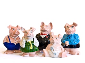 Five ceramic pig figurines are dressed in various outfits. Two pigs wear formal attire, with one in a tuxedo and the other in a business-like shirt and tie. Another pig is dressed in a green dress and yellow tie, and another one in a blue sweater with a bow. The final pig is sitting down, wearing a diaper with a safety pin.