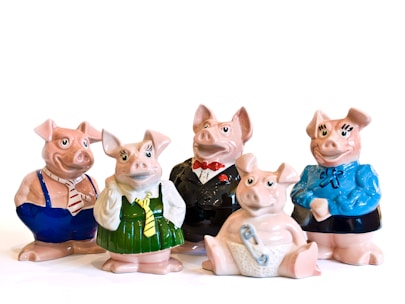 Five ceramic pig figurines are dressed in various outfits. Two pigs wear formal attire, with one in a tuxedo and the other in a business-like shirt and tie. Another pig is dressed in a green dress and yellow tie, and another one in a blue sweater with a bow. The final pig is sitting down, wearing a diaper with a safety pin.