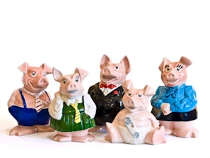 Five ceramic pig figurines are dressed in various outfits. Two pigs wear formal attire, with one in a tuxedo and the other in a business-like shirt and tie. Another pig is dressed in a green dress and yellow tie, and another one in a blue sweater with a bow. The final pig is sitting down, wearing a diaper with a safety pin.