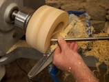 A person is using a wood lathe to shape a piece of wood. The hand is holding a tool that is carving the wood, creating wood shavings that spread around. The lathe is securely holding the wood, spinning it at a constant speed.