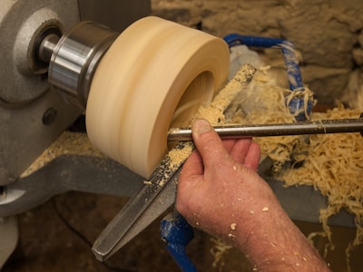 A person is using a wood lathe to shape a piece of wood. The hand is holding a tool that is carving the wood, creating wood shavings that spread around. The lathe is securely holding the wood, spinning it at a constant speed.