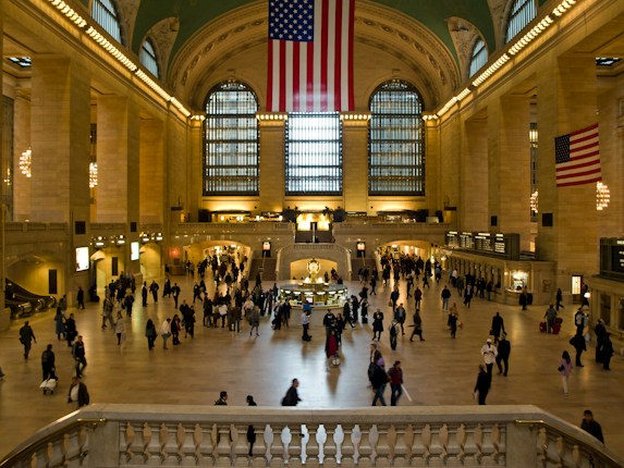A grand, bustling interior of a train station with high arched ceilings, large American flags hanging prominently, and numerous people walking across the spacious concourse. The space is warmly lit, highlighting the architectural details and the grandeur of the hall.