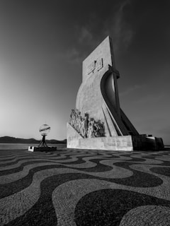 A large monument with detailed sculptures is positioned on a patterned stone pavement, with a spherical structure nearby. The scene is set against a backdrop of a river and distant hills under a clear sky.