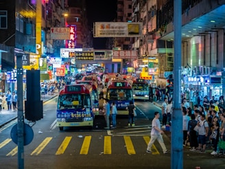 A bustling urban street scene at night illuminated by bright neon signs in various colors. Several mini buses are lined up on the road, surrounded by a crowd of people on the sidewalks. The atmosphere is lively with a multitude of signs advertising different businesses and creating a vibrant glow.