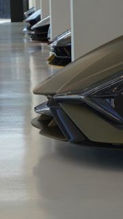 A row of futuristic car front ends are aligned in a showroom or display area. The design features sharp, angular lines and smooth surfaces, reflecting light in a way that emphasizes the modern aesthetic. The floor is polished, adding to the sleek and sophisticated environment.